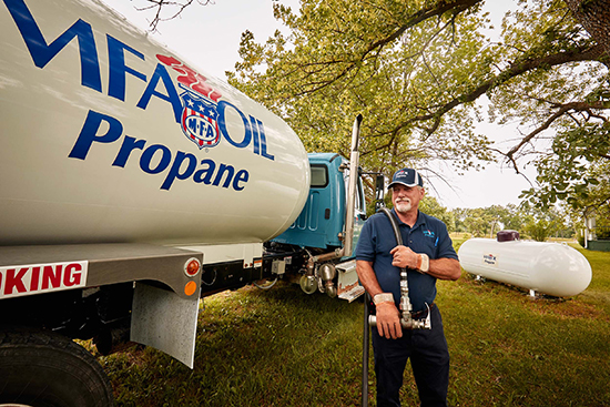 A man in MFA Oil uniform stands beside an MFA Oil propane delivery truck, holding equipment. A propane tank and trees are visible in the background, with the scene set outdoors on a grassy area.