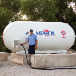 A person in a blue uniform operates a hose connected to a large white propane tank labeled MFA Oil outdoors. The tank is situated on a gravel surface, surrounded by green foliage. Theres a fire extinguisher nearby.