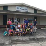 A group of children and adults posing outside a building labeled Welcome to PowerUP! North Missouri Center for Youth & Families. The children are seated in front while the adults stand behind them. Some kids hold papers with drawings.