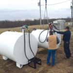 Two men are installing or moving large white fuel tanks in an open field. One man operates machinery, while the other guides the positioning of a tank. The scene is set on a cloudy day with utility poles in the background.