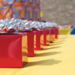 A row of small red gift boxes with silver bows is lined up on a yellow tablecloth. In the background, a person stands near a colorful wall made of interlocking brick toys. The focus is on the boxes, creating a festive atmosphere.