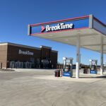Gas station with a convenience store named BreakTime in the background. Several fuel pumps are under a white canopy, and the forecourt is empty. The sky is clear and blue.