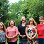 Five women stand outside on a grassy area with trees in the background. They are dressed in casual, colorful attire and are smiling at the camera.