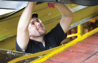A mechanic wearing safety glasses and a cap is working underneath a vehicle, looking up with a focused expression. He is reaching up with red gloves, surrounded by industrial equipment in a workshop setting.