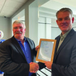 Two men in suits are shaking hands. One is holding a framed certificate. A vintage gas pump is visible in the background.