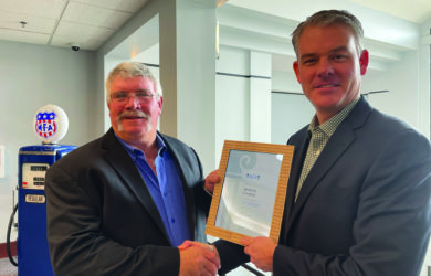 Two men in suits are shaking hands. One is holding a framed certificate. A vintage gas pump is visible in the background.