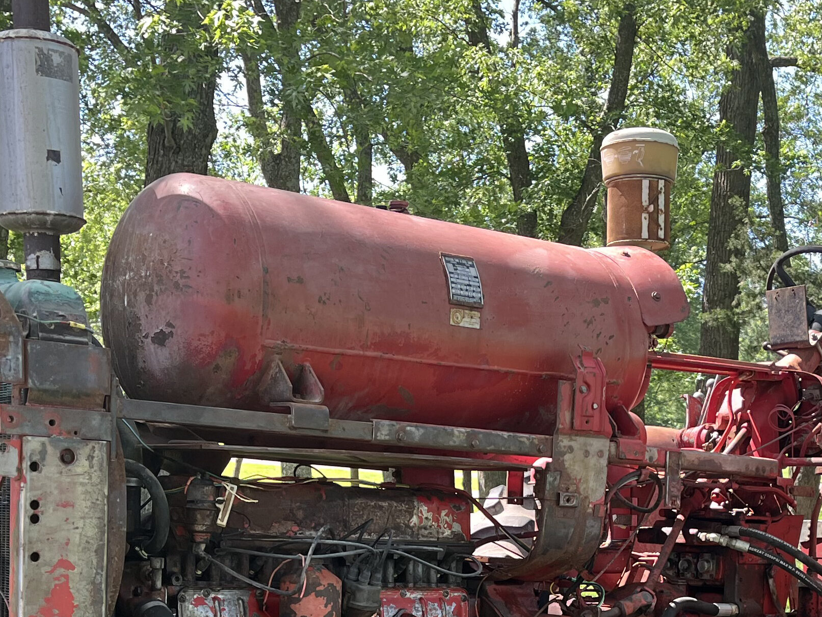A rusty red vintage tractor sits on green grass under tall trees in daylight. In the background, part of a green tractor is visible among the shade and trees.