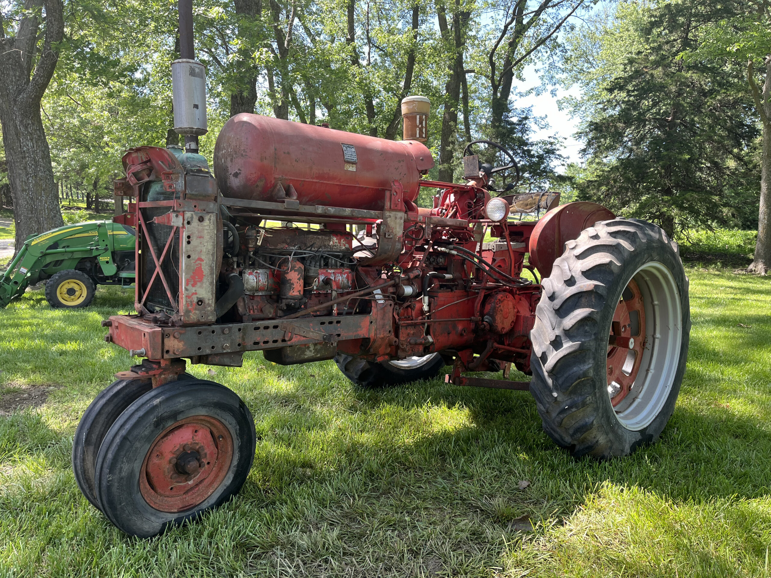 A rusty red vintage tractor sits on green grass under tall trees in daylight. In the background, part of a green tractor is visible among the shade and trees.