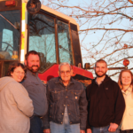 A group of five people smiles in front of a red tractor during sunset. Bare tree branches frame the background, creating a warm, peaceful atmosphere.