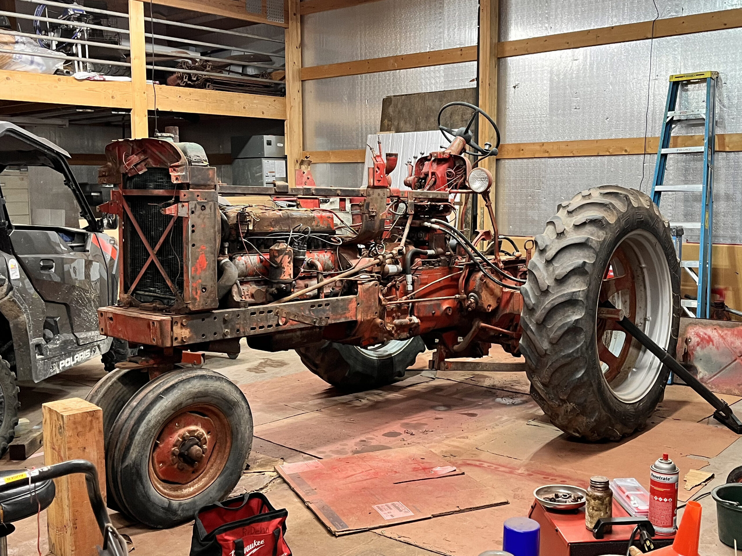 A red, partially disassembled tractor sits in a cluttered workshop with tools, parts, and equipment scattered around. An off-road vehicle and a ladder are also visible.