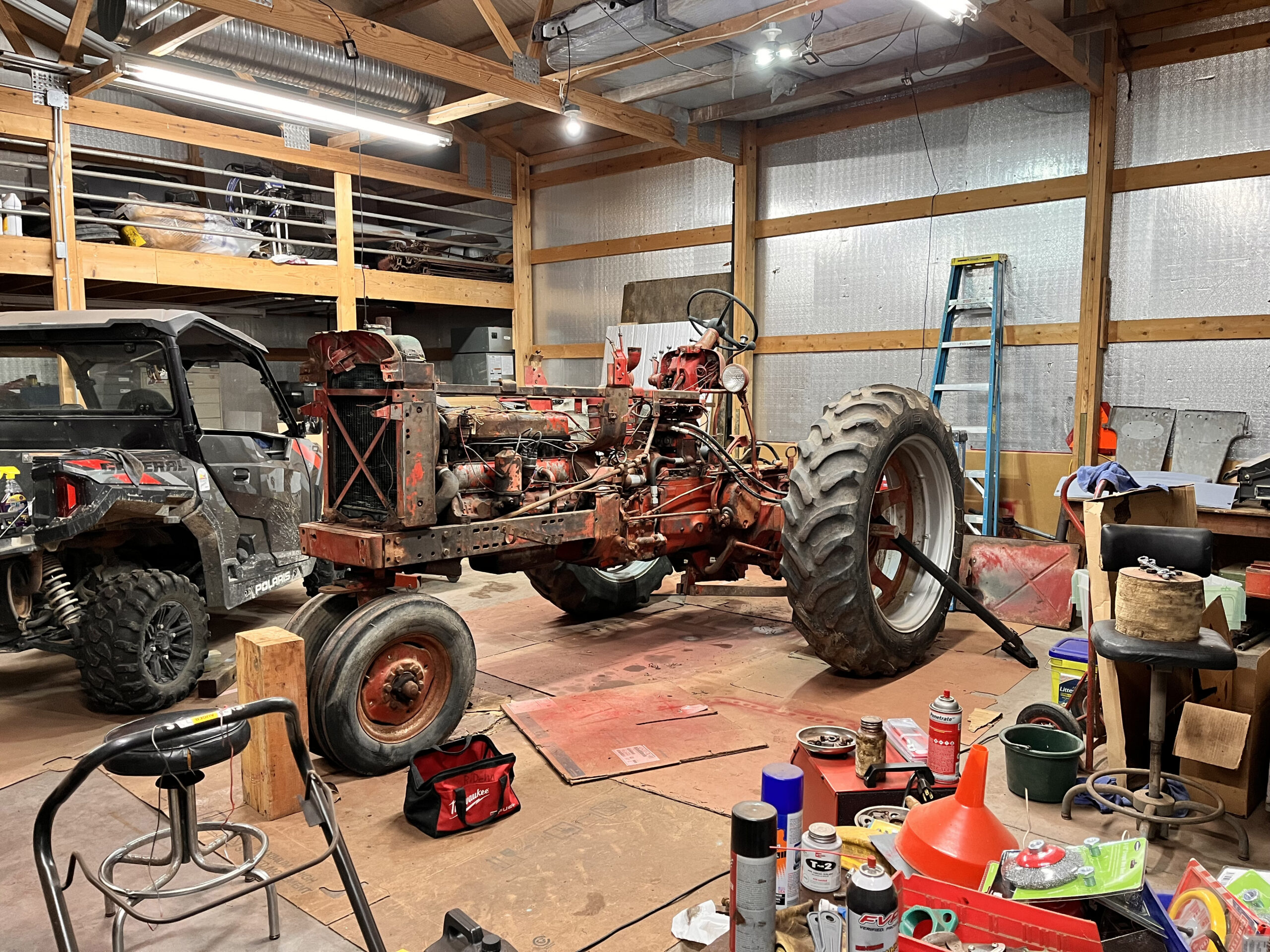A red, partially disassembled tractor sits in a cluttered workshop with tools, parts, and equipment scattered around. An off-road vehicle and a ladder are also visible.