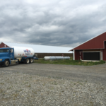 A blue fuel truck is parked on a gravel lot beside a large red barn. The sky is overcast with gray clouds, and there are propane tanks near the building.
