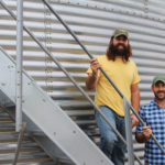 Two men stand on metal stairs in front of a large corrugated metal structure. One wears a yellow shirt and jeans with a cap, the other a blue plaid shirt and cap. They are both smiling.
