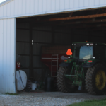 A large metal barn with an open sliding door reveals a green tractor parked inside. An above-ground white fuel tank with a pump is situated outside on a gravel area. A smaller building with a metal roof is visible in the background.