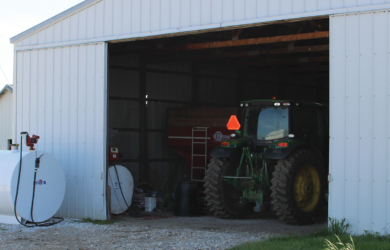 A large metal barn with an open sliding door reveals a green tractor parked inside. An above-ground white fuel tank with a pump is situated outside on a gravel area. A smaller building with a metal roof is visible in the background.