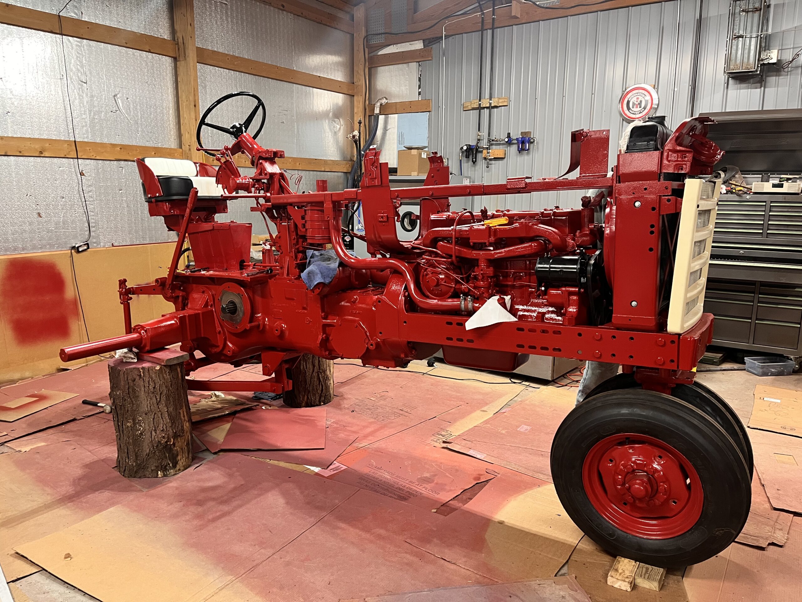 A partially restored red tractor with missing panels and parts sits indoors on tree stumps, surrounded by tools, spray paint, and cardboard sheets in a workshop.