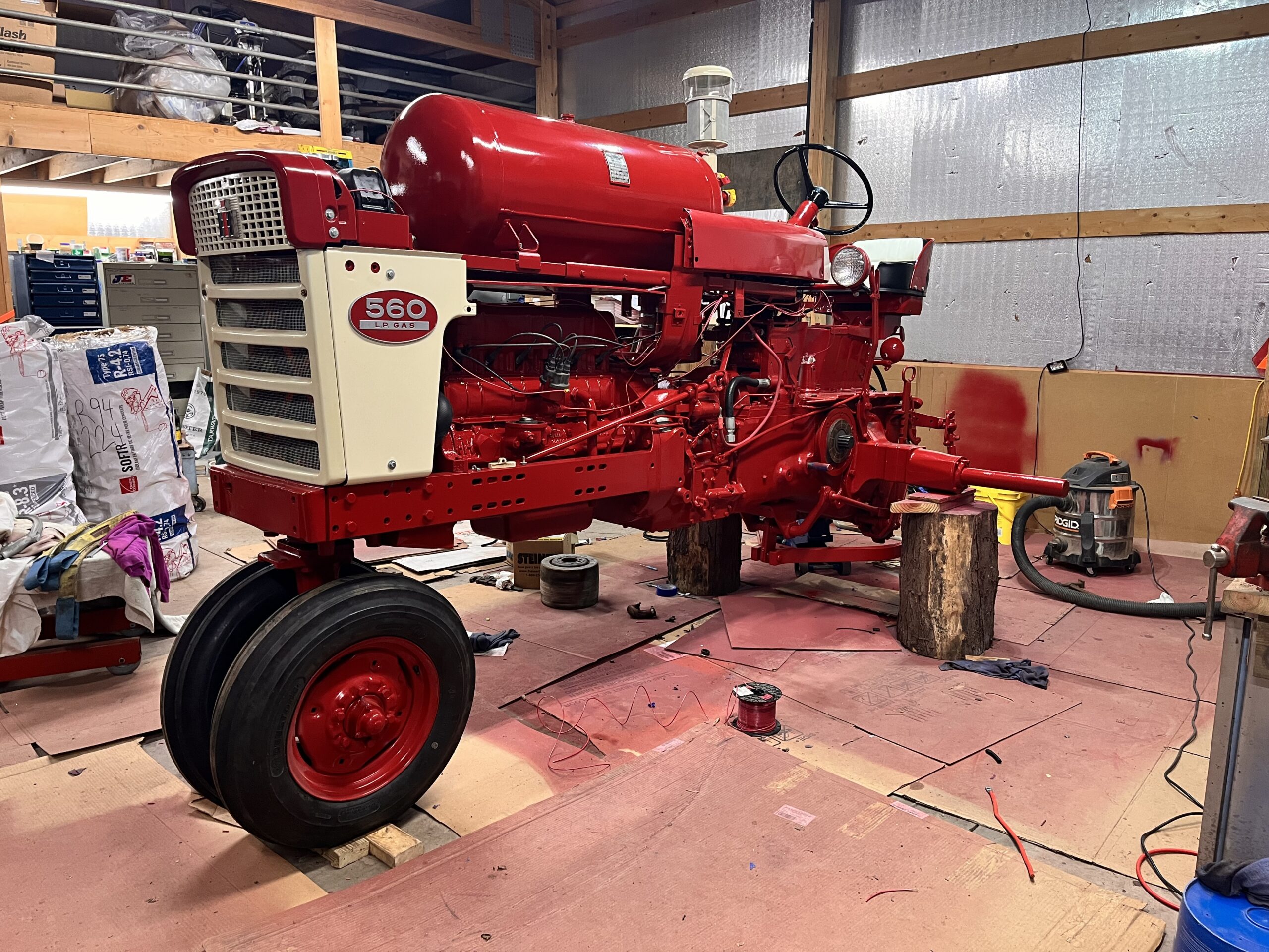 A red vintage Farmall 560 tractor is being restored in a workshop, with the front wheels attached and the back raised on wooden blocks. Paint supplies and tools are scattered on the floor.