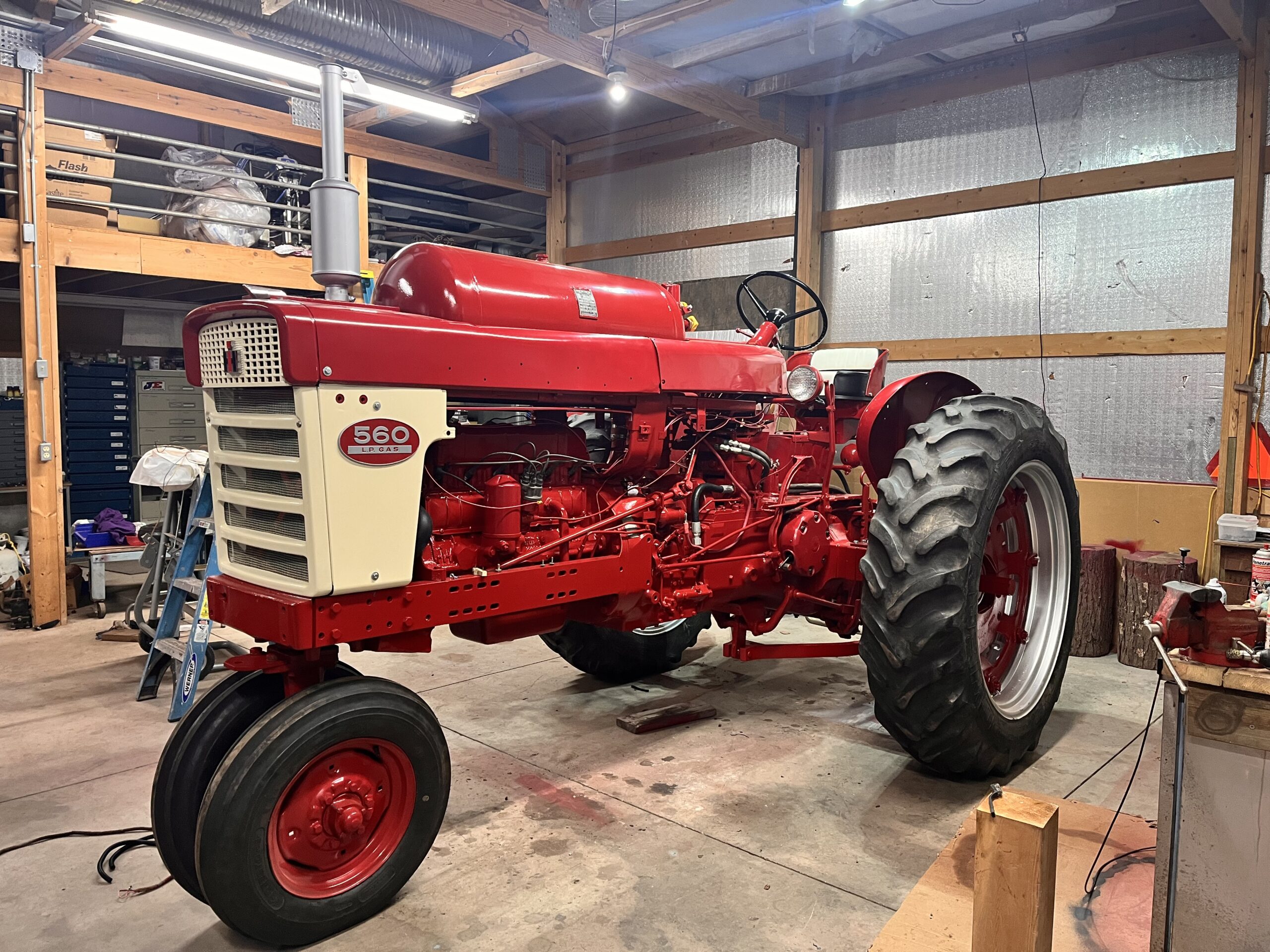 A restored red and white vintage Farmall 560 tractor sits inside a well-lit garage with tools, shelves, and wooden beams visible in the background.