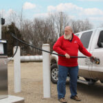 A bearded man in a red sweater is filling a white truck with propane auto gas at a fueling station. The background shows trees and several large propane tanks.