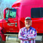 A man with a white beard and a cap stands with arms crossed in front of a red semi-truck. He is wearing a plaid shirt. Trees are visible in the background.
