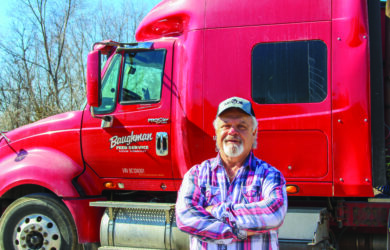 A man with a white beard and a cap stands with arms crossed in front of a red semi-truck. He is wearing a plaid shirt. Trees are visible in the background.