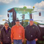 Three people stand in front of a large green tractor under a partly cloudy sky. They wear casual clothing, including hats and hoodies, and are smiling at the camera. The Gillilands appear ready for another day on the farm, embracing both work and camaraderie in their rustic setting.