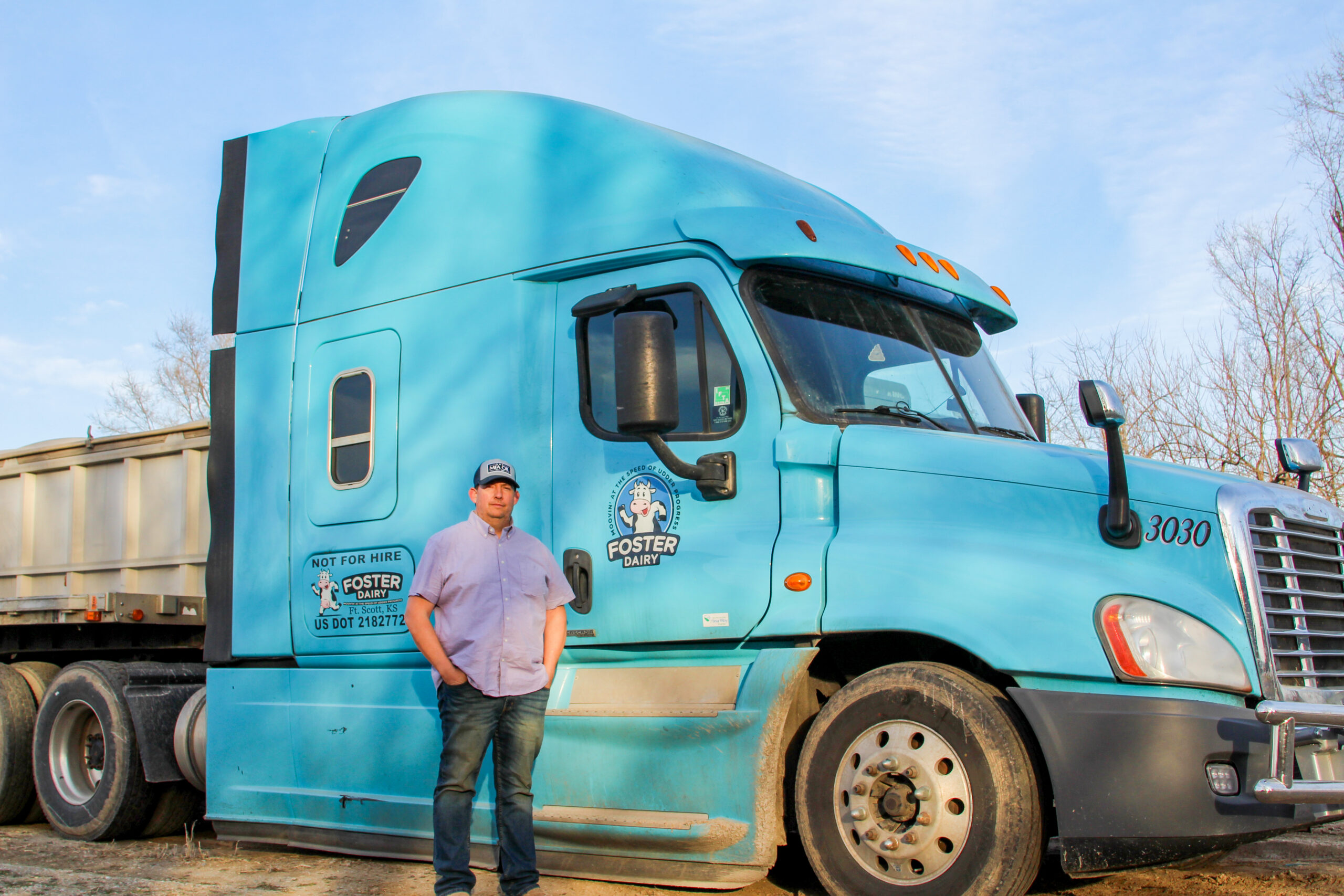 A man in a cap and jeans stands in front of a large blue semi-truck with “Foster Dairy” branding, parked outdoors on a clear day. Trees and part of a trailer are visible in the background.