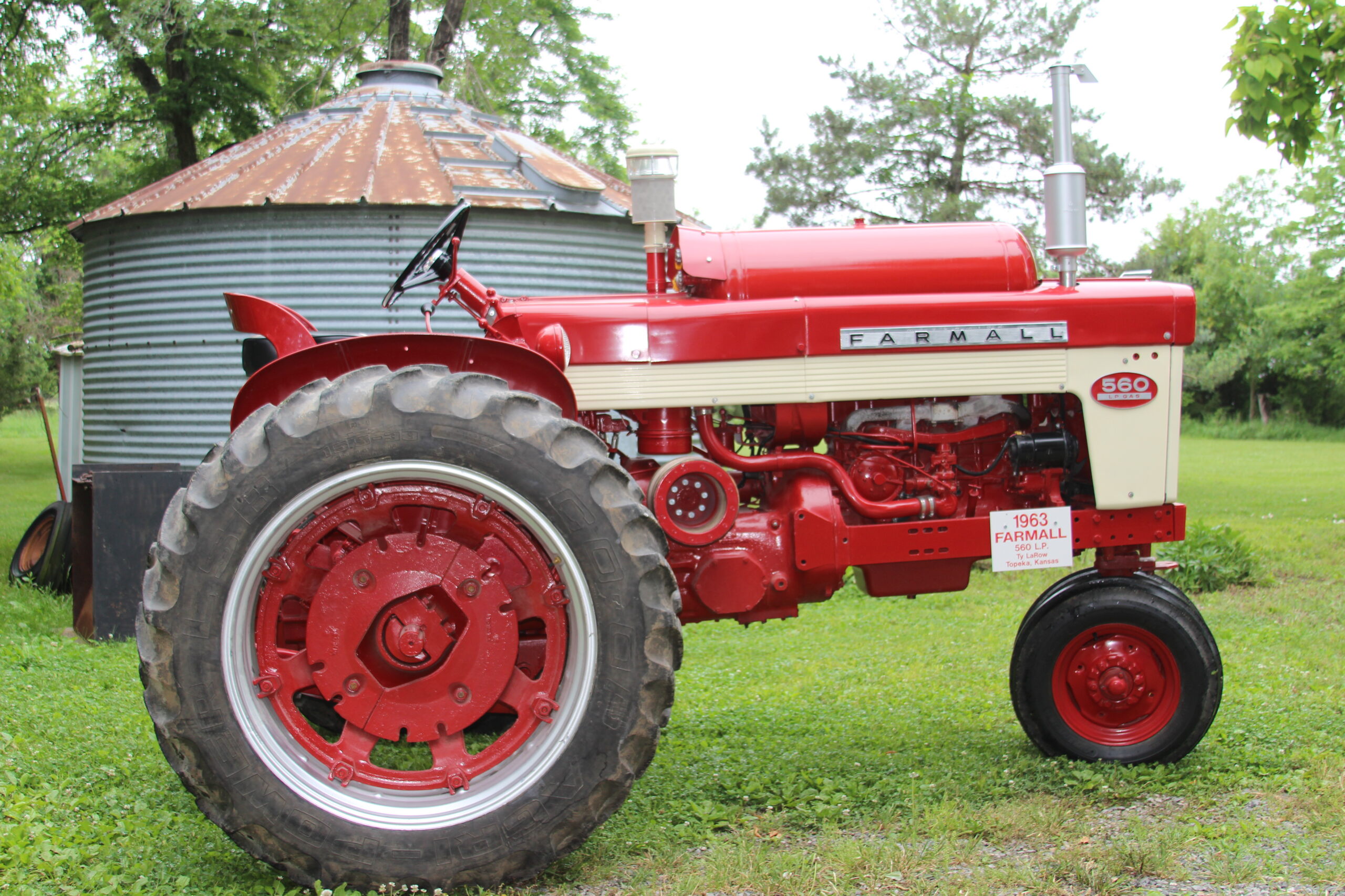 A red and white Farmall 560 tractor is parked on grass in front of a round, metal grain bin and trees. The tractor is clean and appears restored.