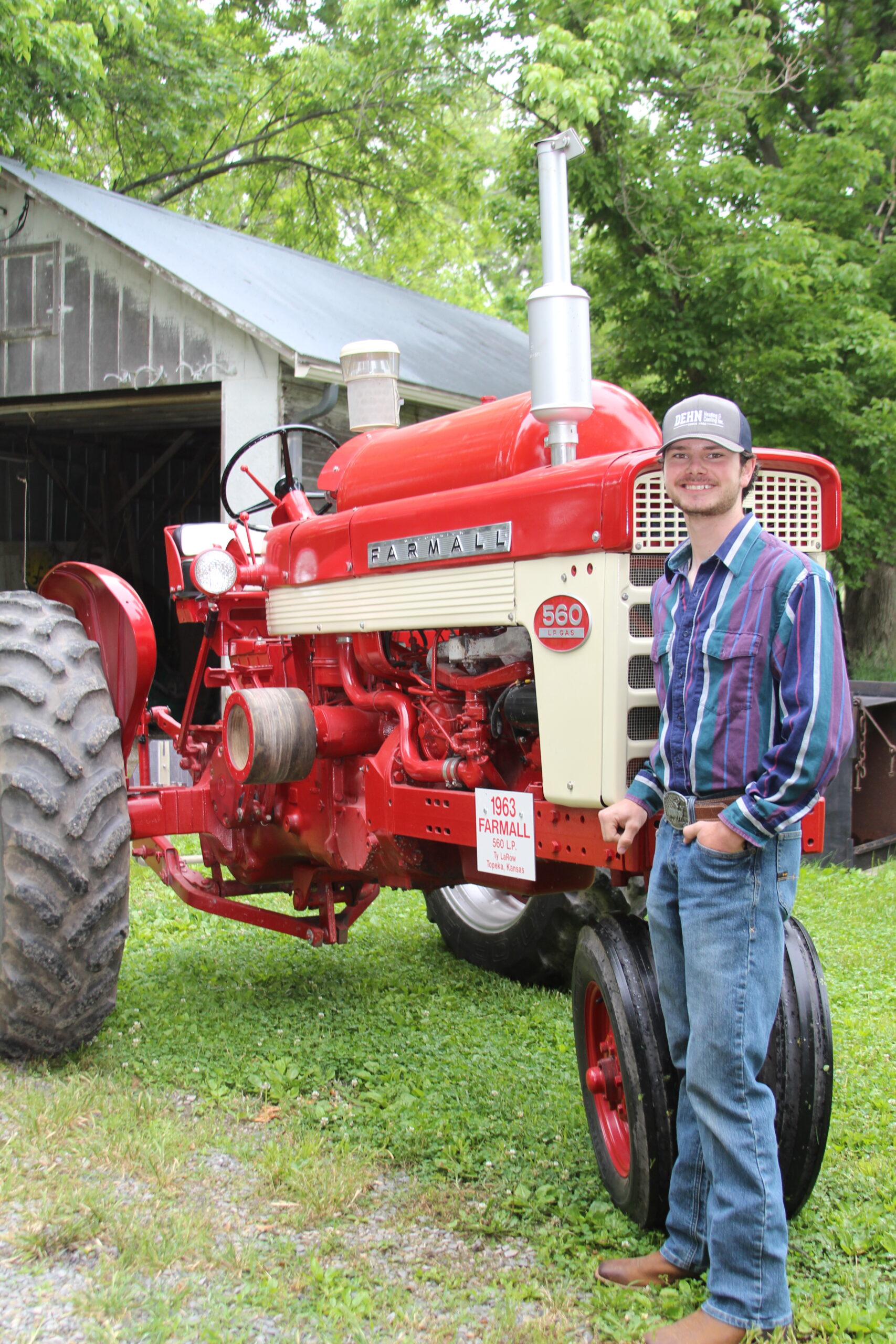 Ty La Row in a striped shirt and jeans stands smiling next to a restored red Farmall 560 tractor in front of a barn with green trees in the background.