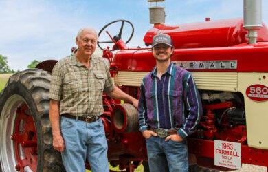 Two people stand beside a red Farmall 560 tractor on a farm under a blue sky. One person wears a plaid shirt and jeans; the other wears a striped shirt, jeans, and a hat. A grassy field is visible in the background.