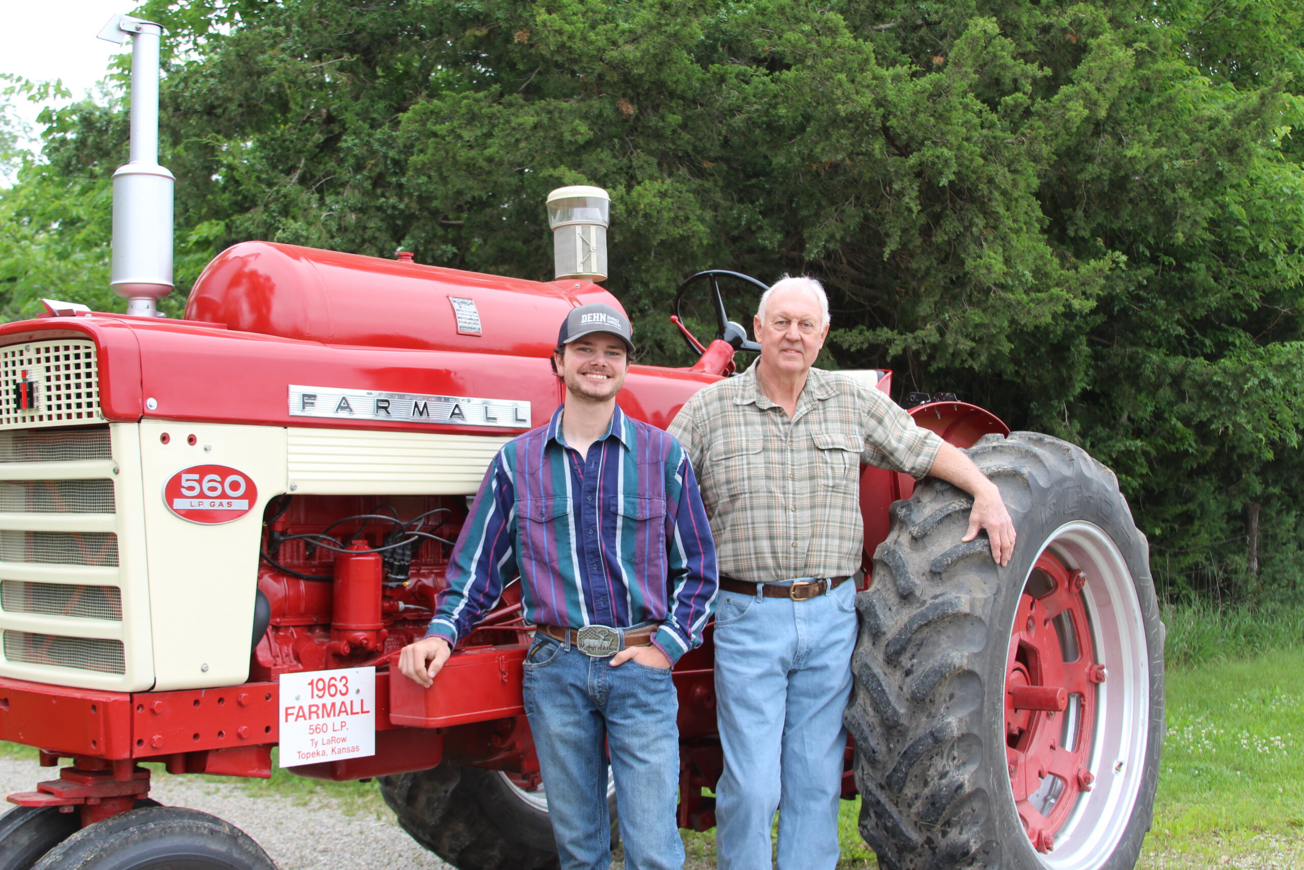 Ty La Row stands next to a vintage red Farmall tractor outdoors. The younger man wears a striped shirt and cowboy hat; the older man wears a plaid shirt. Green trees fill the background.