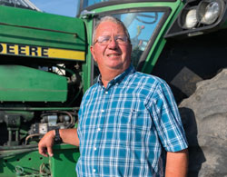A man in a blue plaid shirt stands smiling beside a green tractor under a clear blue sky.