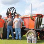 Three men stand in front of a large orange Allis-Chalmers tractor on grass. The tractor is adorned with a horse logo and the number 21. Two containers are placed near the tractors front wheel.