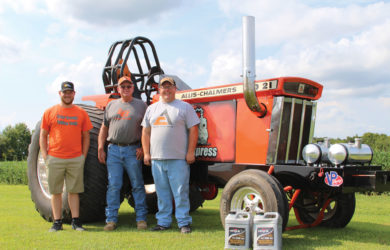 Three men stand in front of a large orange Allis-Chalmers tractor on grass. The tractor is adorned with a horse logo and the number 21. Two containers are placed near the tractors front wheel.
