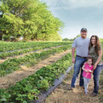 A family of three stands smiling in a green field with rows of plants. They are surrounded by trees under a clear blue sky. The man wears a cap, the woman has long hair, and the child is holding a small plastic basket.