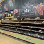An almost empty supermarket meat section with a few packages remaining. Signs advertise natural beef options. A digital sign shows the price of ground beef as $9.48. The shelves are mostly bare, indicating low stock.