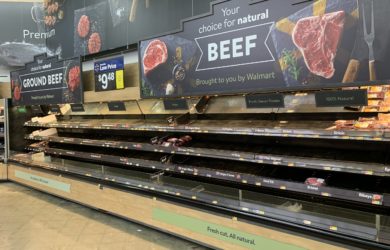 An almost empty supermarket meat section with a few packages remaining. Signs advertise natural beef options. A digital sign shows the price of ground beef as $9.48. The shelves are mostly bare, indicating low stock.