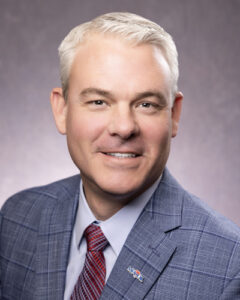 Portrait of Jon Ihler President and Chief Executive officer of MFA Oil with short gray hair is smiling, wearing a blue checkered suit, a white shirt, and a red striped tie. A lapel pin is visible on the suit. The background is a simple gray gradient.