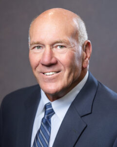 A bald man in a suit and striped tie smiles at the camera against a plain, neutral background.