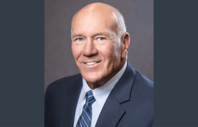 A smiling older man with a bald head and light eyes wearing a dark suit, light blue shirt, and striped tie, posing in front of a plain grey background.