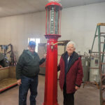 John A. Busch and his wife, Marie, stand indoors beside a tall, vintage red gas pump with an MFA logo and a glass cylinder. The room has workshop tools and equipment. John wears a cap and jacket, and Marie wears glasses and a red coat.