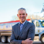 A person in a gray suit smiles confidently with arms crossed, standing outdoors in front of a large truck. The sky is clear and the trees show autumn foliage. The truck is white and blue with red accents.