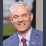 MFA Oil President & CEO Jon Ihler wears a blue checked suit, light blue shirt, and red striped tie. He stands in front of a blurred outdoor background.