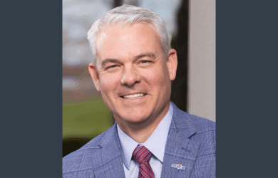 MFA Oil President & CEO Jon Ihler wears a blue checked suit, light blue shirt, and red striped tie. He stands in front of a blurred outdoor background.