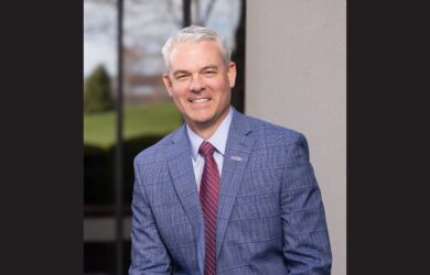 MFA Oil President and CEO Jon Ihler in a blue plaid suit, white shirt, and red striped tie smiles while posing outdoors in front of a building with large windows and a concrete wall.