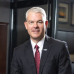 A person with short gray hair, exuding trust, stands indoors wearing a dark suit, white shirt, and red striped tie. They smile warmly at the camera, surrounded by framed pictures and a counter in the background.