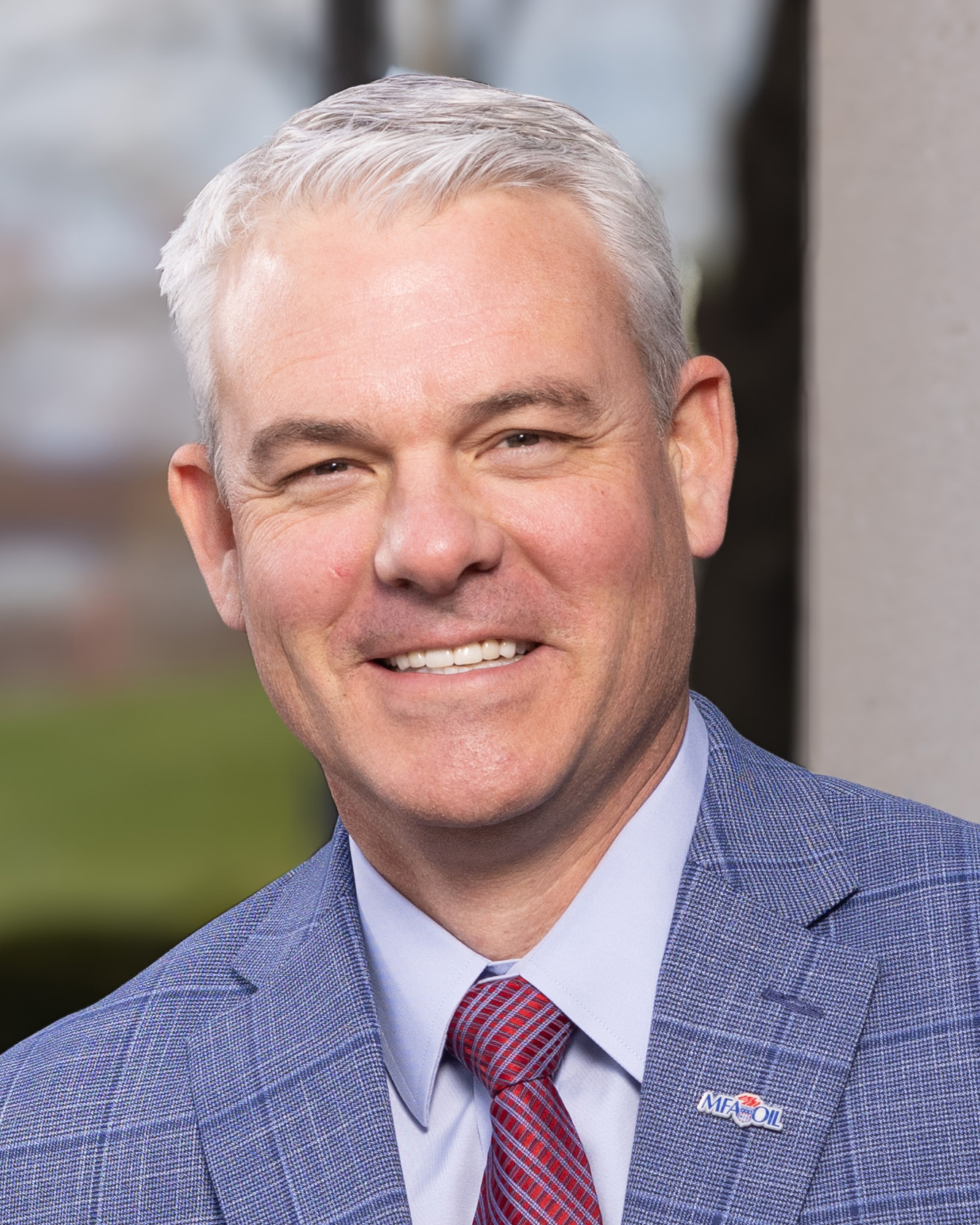 MFA OIl President & CEO wears a blue checked suit, light blue shirt, and red striped tie. He stands in front of a blurred outdoor background. 