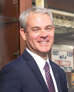 Jon Ihler, MFA Oil president and CEO, is smiling, wearing a navy suit, white shirt, and striped tie. Behind him are vintage MFA Oil signs, displayed in a wooden cabinet.