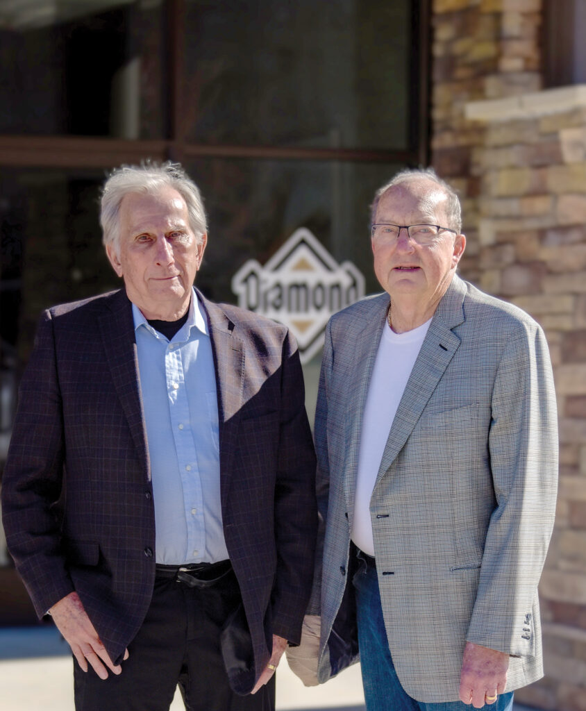 Richard Kampeter & Gary Schell stand outside in front of a building with a stone facade and a glass door. Both wear blazers; one is in a dark jacket with a blue shirt, the other in a gray jacket with a white shirt and jeans.