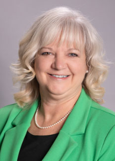 A smiling woman with wavy, shoulder-length gray hair, wearing a green blazer over a black top and a pearl necklace, posed against a plain gray background.